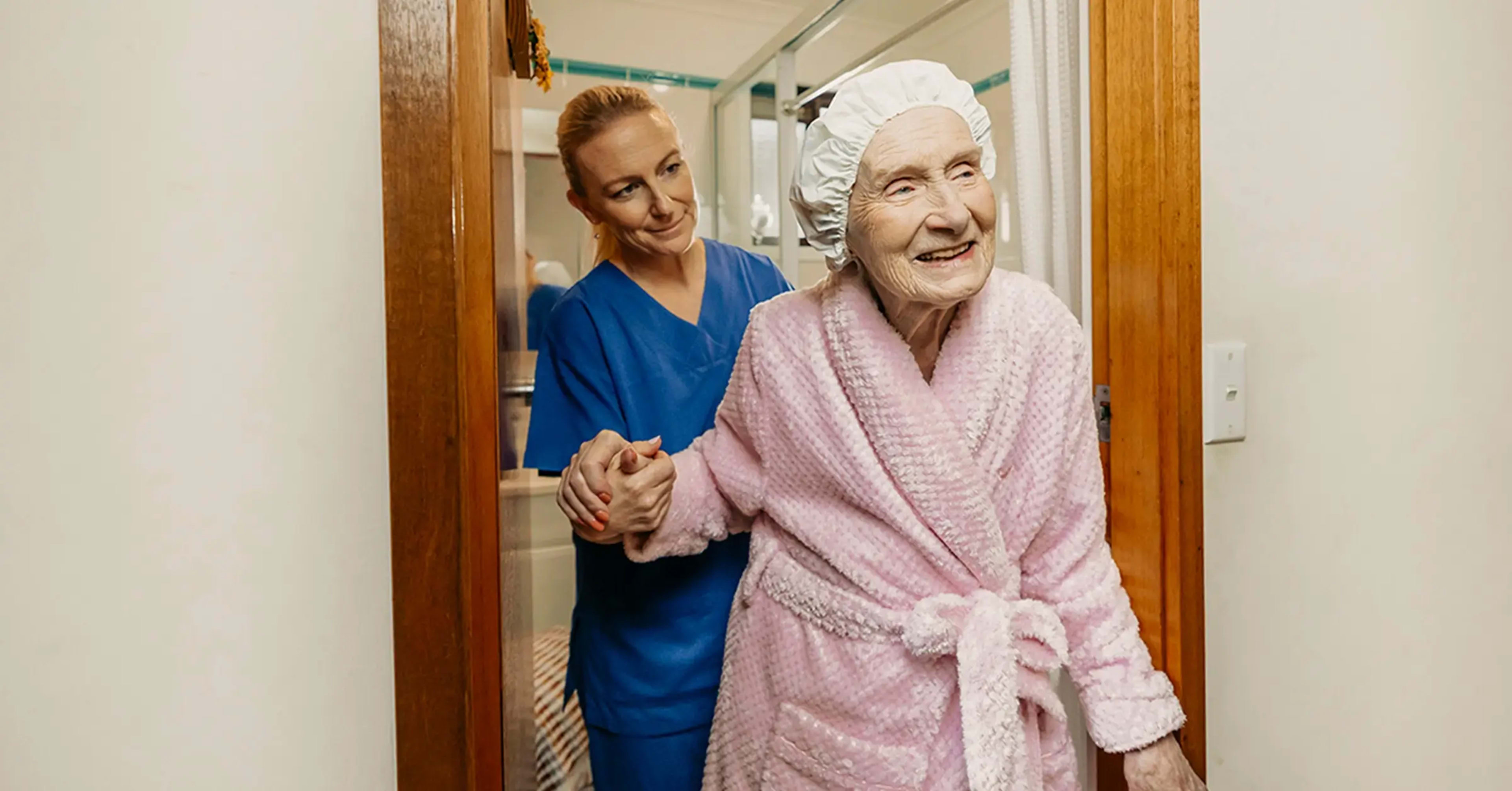 Registered Nurse in scrubs helps older client in pink bathrobe walk out of their bathroom.