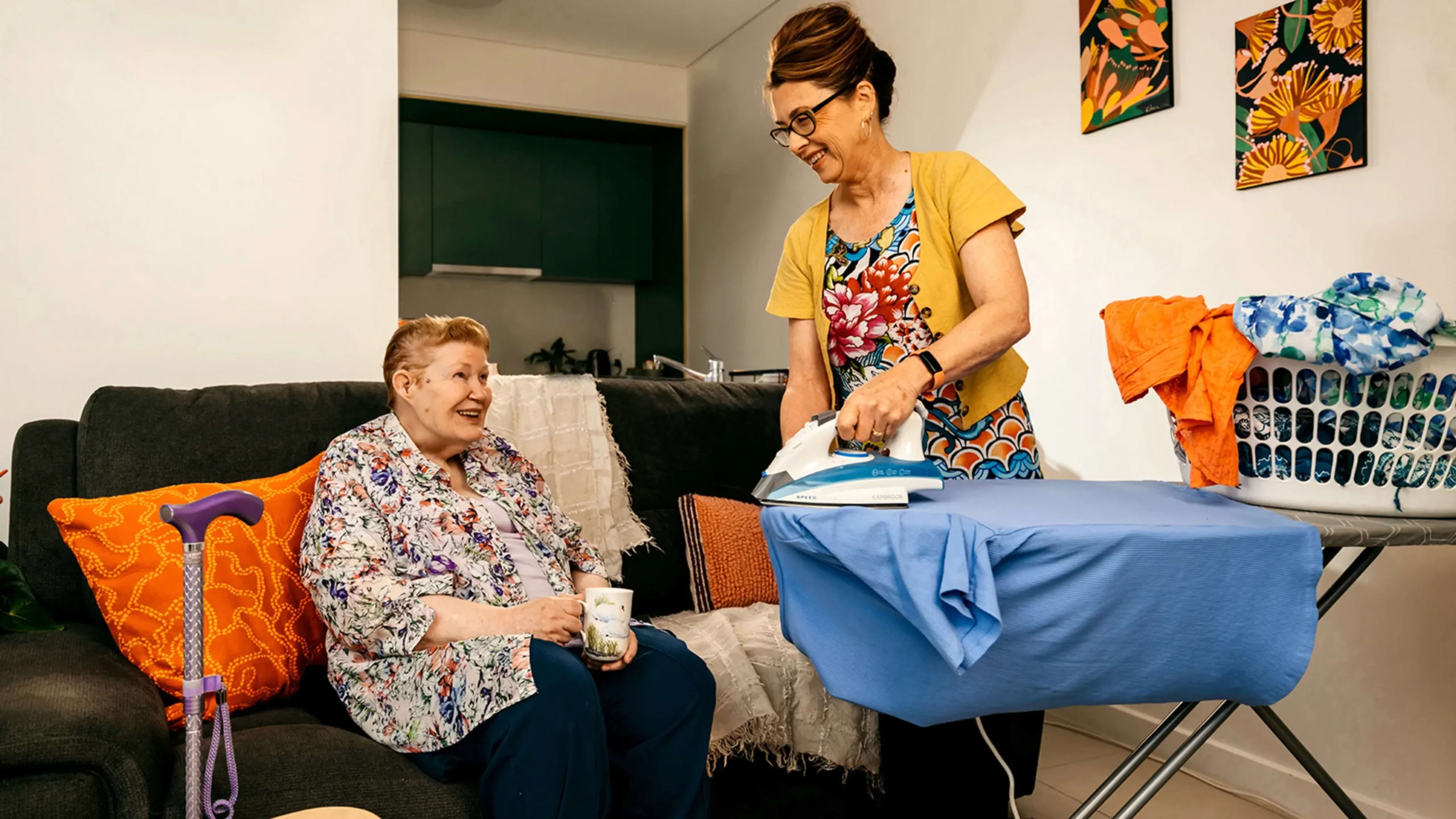 Older person with a walking stick sits on the couch with tea and chats to support worker ironing.