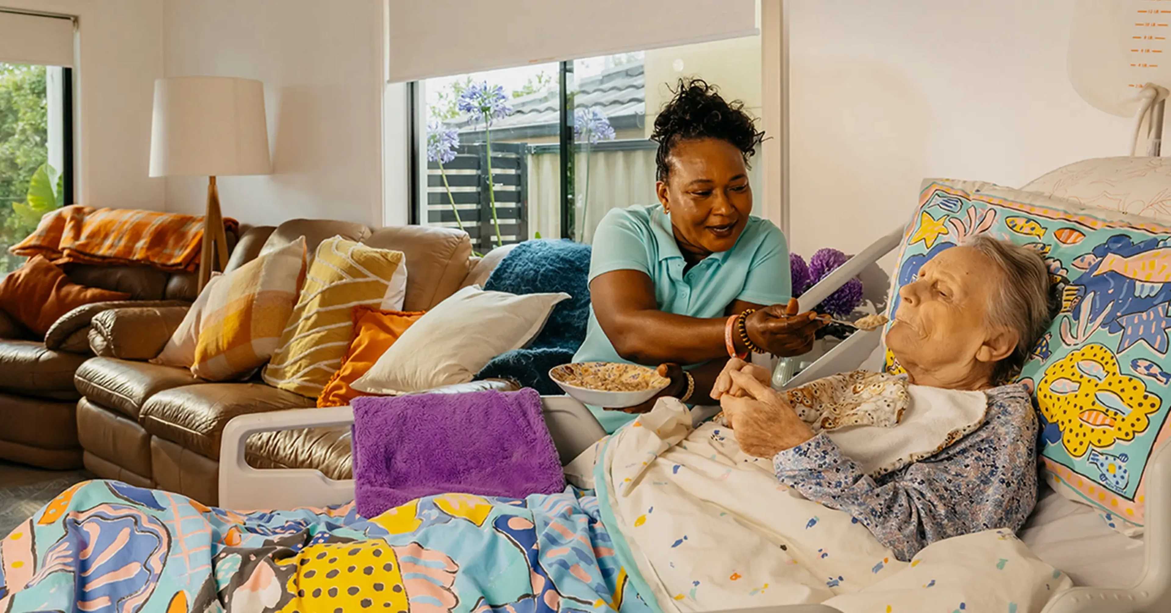 Support worker smiles as they feed an older person living with Dementia laying in bed at home.