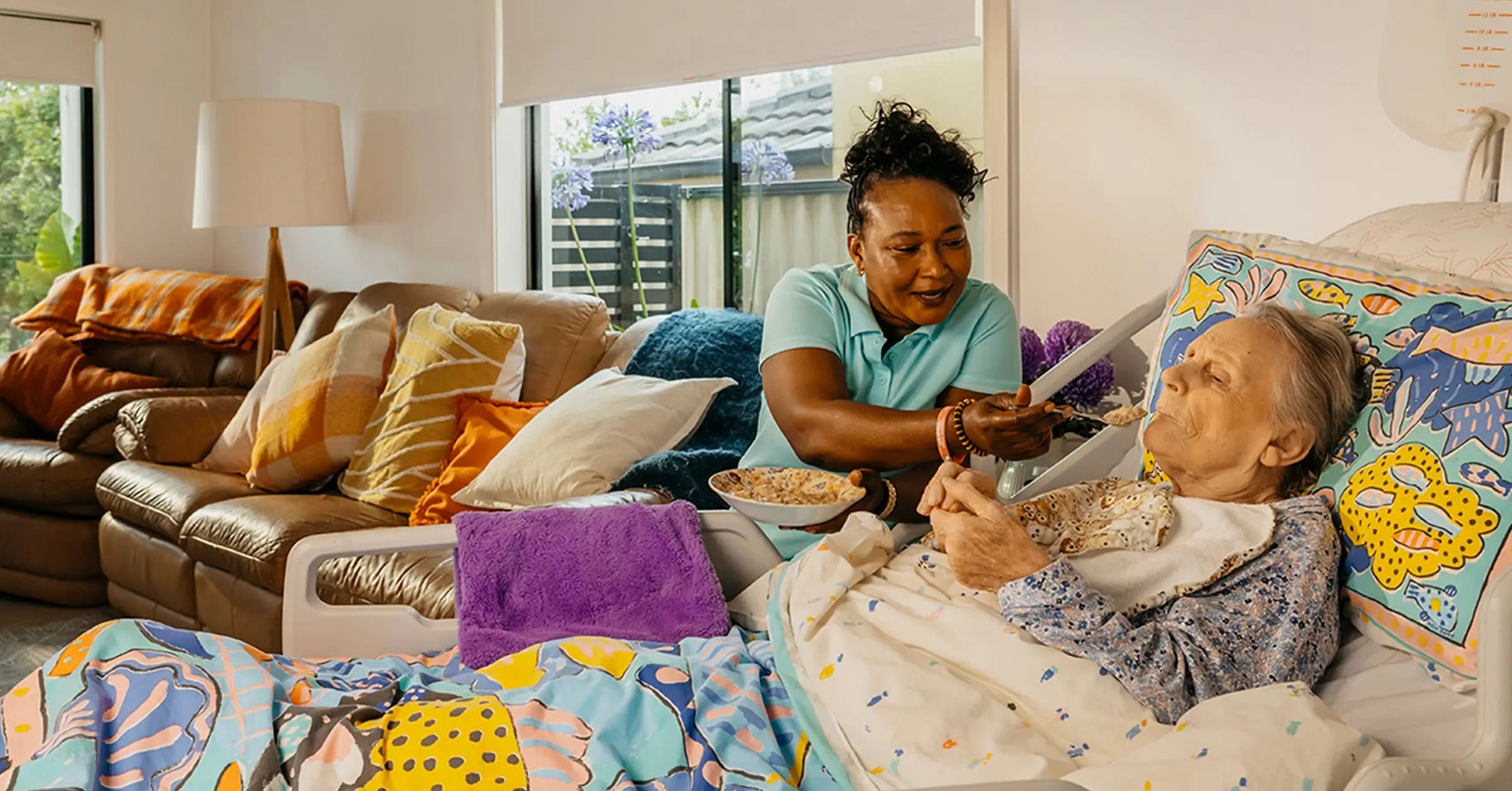 Support worker smiles as they feed an older person living with Dementia laying in bed at home.