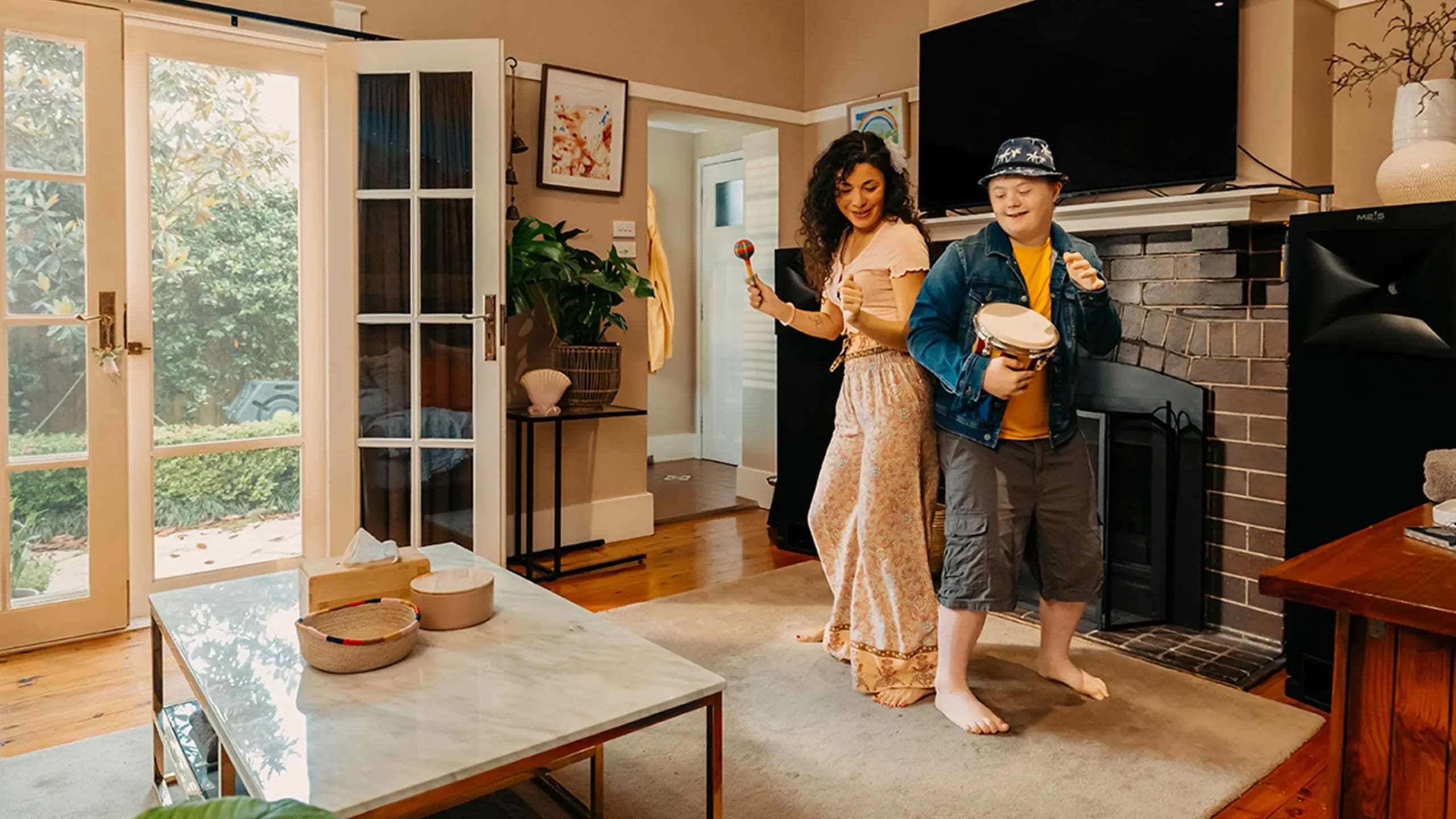Child with disability beats drum and support worker shakes maraca as they dance in living room.