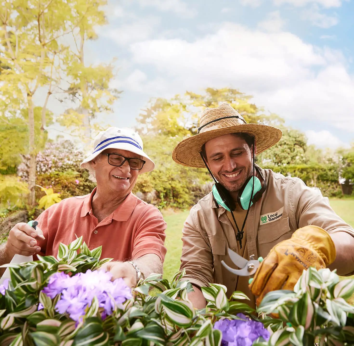 An older man and a smiling gardener prune flowering plants together in a sunny garden.